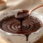 A mason jar of Homemade All Natural Chocolate Syrup next to a fresh glass of chocolate milk and a whisk.